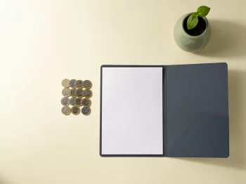 Stapel euromunten naast een open documentmap en een groen plantje in keramieken pot op een minimalistisch bureau.