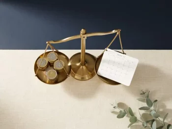 Messing weegschaal met euromunten en maandkalender op linnen ondergrond, bovenaanzicht, minimalistische compositie.