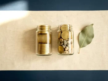 Twee glazen potjes met goudkleurige munten op linnen ondergrond, één pot gemengd met kiezelstenen als symbool voor passief beleggen.