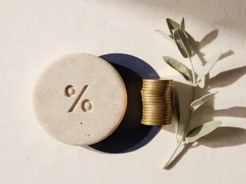 Percentage-symbool gegraveerd in stenen schijf naast stapel gouden munten op crème linnen ondergrond.