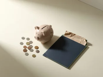 Gekanteld keramisch spaarvarken met verspreide munten en een vervagende documentmap op een crèmekleurig bureau.