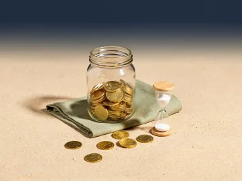 Halfgevuld glazen pot met gouden munten naast een zandloper op zandkleurig oppervlak, marineblauw-crème achtergrond.