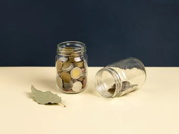 Glazen pot gevuld met munten naast een omgevallen lege pot op een crème ondergrond met marineblauw achtergrond.