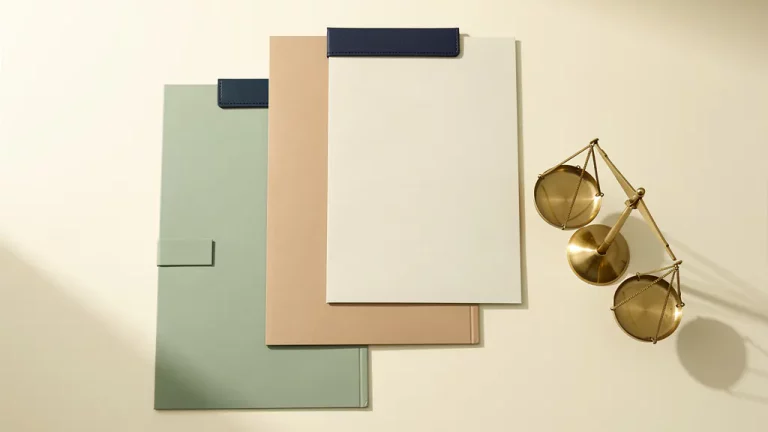 Drie geordende documentmappen op een crèmekleurig bureau naast een messing weegschaal, gestyled in salie groen en marineblauw.