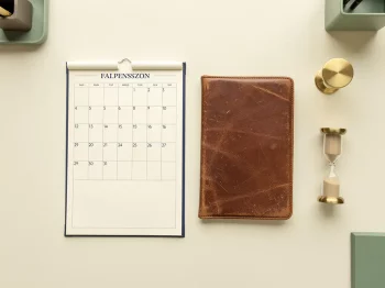 Vintage Nederlandse kalender naast een versleten leren pensioendossier en messing zandloper op een crèmekleurig bureau.