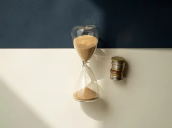 Zandloper met stromend zand naast een stapel munten op een crèmekleurig oppervlak, bovenaanzicht in zacht natuurlijk licht.